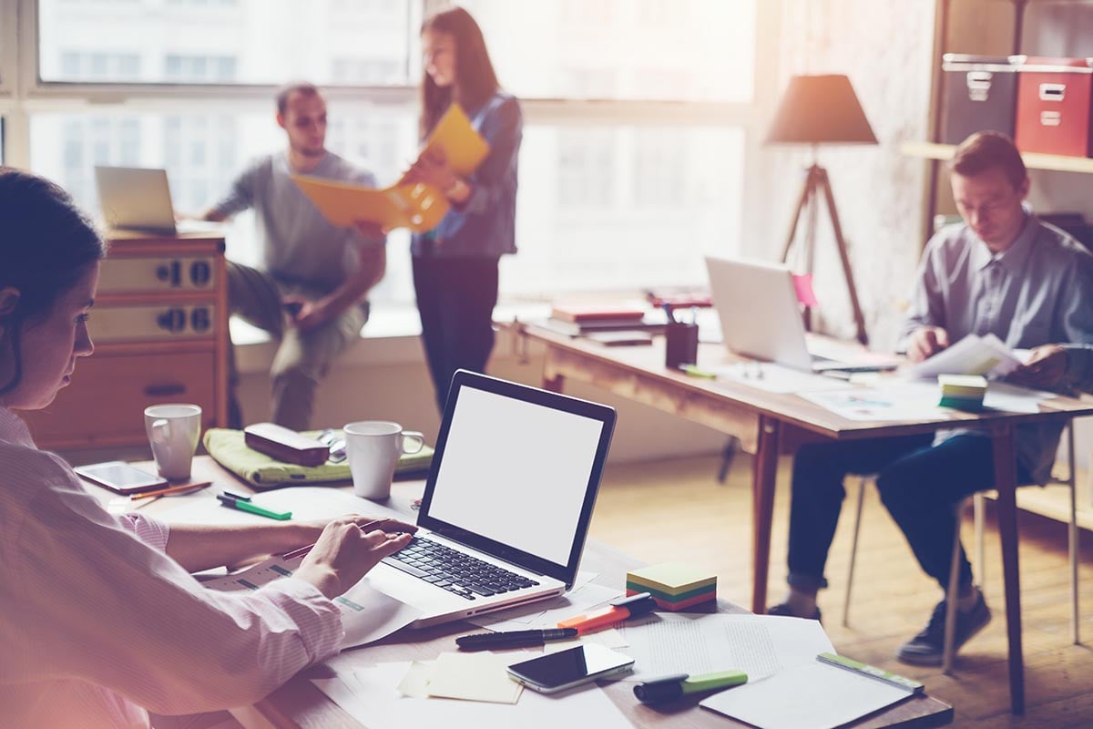 Symbolfoto für Microsoft 365 - Büro mit mehreren Schreibtischen und Computern an den 4 Personen sitzen bzw. stehen