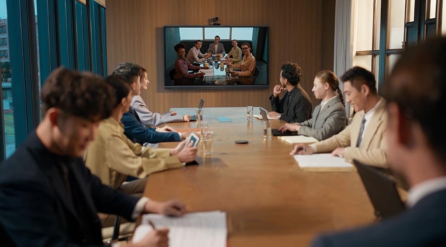Darstellung einer Videokonferenz. Am Besprechungstisch sitzen 9 Personen auf dem Display an der Wand sind 5 Personen zu sehen. Das Bild soll die Audioqualität (Lautsprecher und Mikros) des MeetingBoards Pro darstellen.