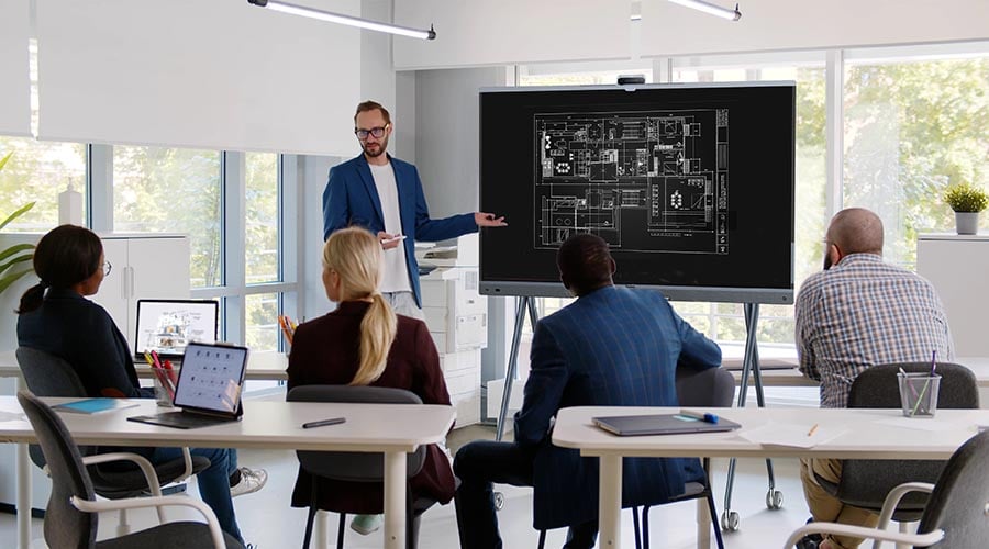 Auf dem Bild wird eine Schulung dargestellt. Der Moderator steht am MeetingBoard Pro von Yealink und erklärt einen Grundrissplan auf dem Display. Vor dem Display sitzen 4 Personen.