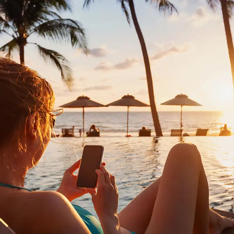 Frau mit Mobiltelefon in der Hand im Urlaub am Pool sitzend. Im Hintergrund sind Palmen, Sonnenschirme und das Meer zu sehen